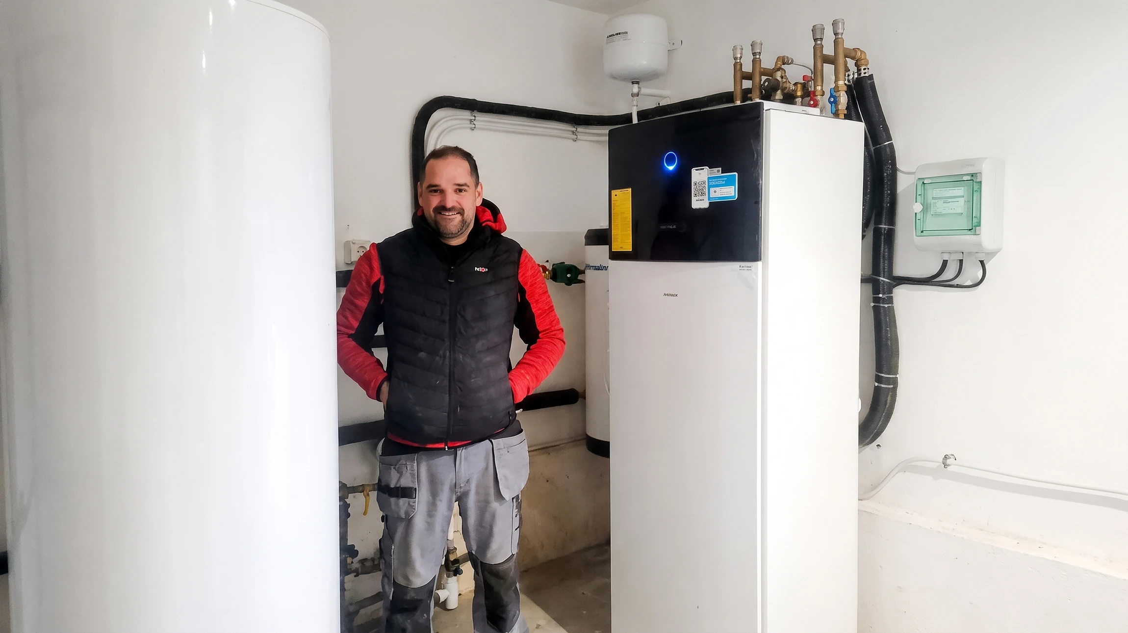 Homme technicien barbu en salopette bleue et t-shirt blanc installe ou répare un chauffe-eau électrique mural blanc dans une cuisine moderne